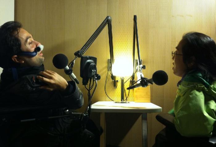 Image of two people in a recording booth with microphones angled near their faces. On the left is a Latino man in a wheelchair using a nasal mask for respiratory support. On the right is an Asian woman in a wheelchair with glasses. They are in conversation with one another.