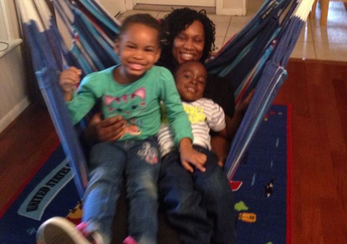 Black woman sitting back in a hammock with two young black boys on her lap. They are all smiling at the camera.