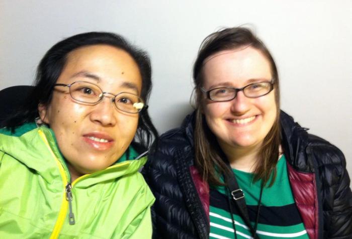 Image of two women sitting side by side. On the left is an Asian woman with glasses and black hair. She is wearing a green jacket. The woman on the right is white and she has long brown hair and glasses. She is wearing a black puffy winter coat and a green sweater.