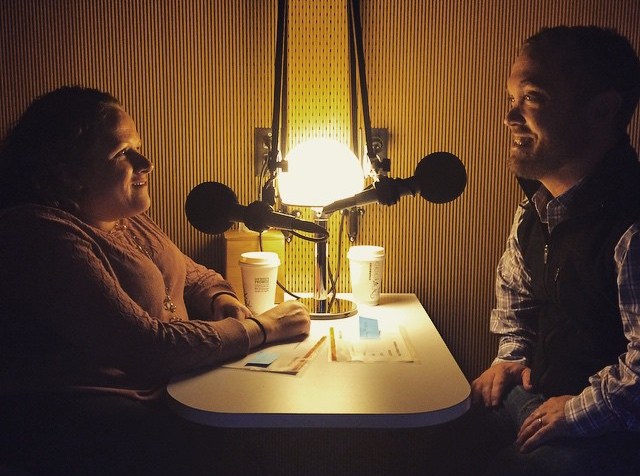 A room with dimmed lighting, two people are sitting across from one another with microphones in front of them. On the left is a middle-aged white woman in a wheelchair with long brown hair. On her right, facing her, is a middle-aged white man with short brown hair.