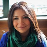 Latina with dark brown hair wearing a blue sweater and a green and black scarf. She is smiling at the camera.