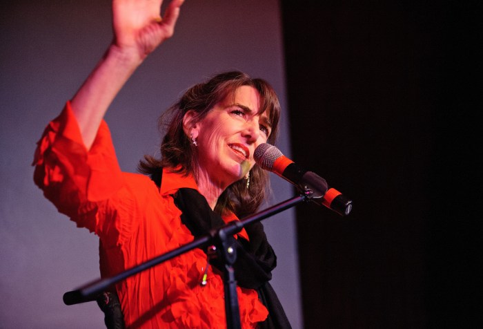 A white woman in a red shirt standing in front of a microphone. Her hand is in the air and looking at something upward.