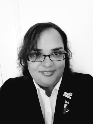 Black and white frontal headshot picture of a Latina woman in her mid to late 20s. She is smiling, has eyeglasses and is wearing a white shirt with a black blazer. In her lapel she is wearing a pin with the logo of the National LGBTQ Task Force, and a brooch in the shape of an owl. 