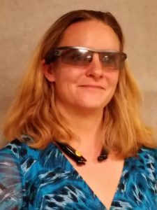 Photo description: A middle aged white woman smiling, as she receives visual interpretation from a remote human guide who gets a bird’s eye view via the camera on her AirA sunglasses. There is a Bluetooth receiver on her neck.