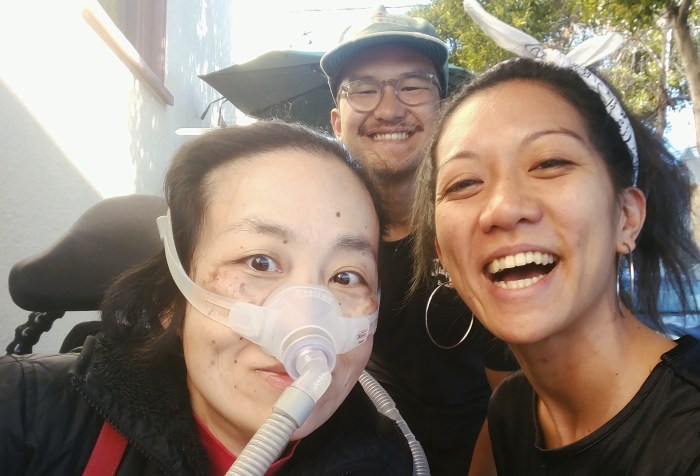 Three Asian Americans taking a selfie at an outdoor sidewalk. In the back is Luigi Villanueva, on the left is Alice Wong, a wheelchair user with a mask over her nose attached to a ventilator, on the right is Rochelle Kwan. Everyone is smiling.