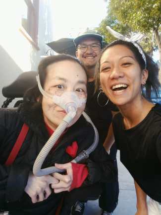 Three Asian Americans taking a selfie at an outdoor sidewalk. In the back is Luigi Villanueva, on the left is Alice Wong, a wheelchair user with a mask over her nose attached to a ventilator, on the right is Rochelle Kwan. Everyone is smiling.