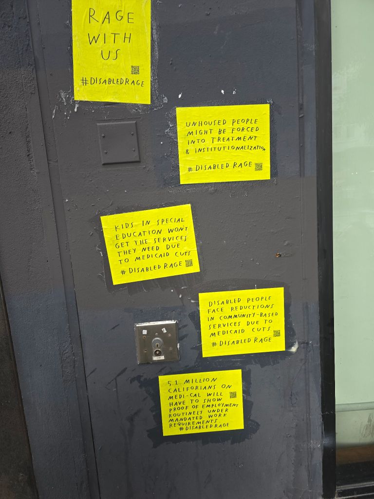 On a wall on the street are 4 pieces of bright yellow paper arranged vertically with various statements all with a QR code and #DisabledRage. One says “RAGE WITH US” while others are about unhoused people, kids in special education, and Medicaid cuts
