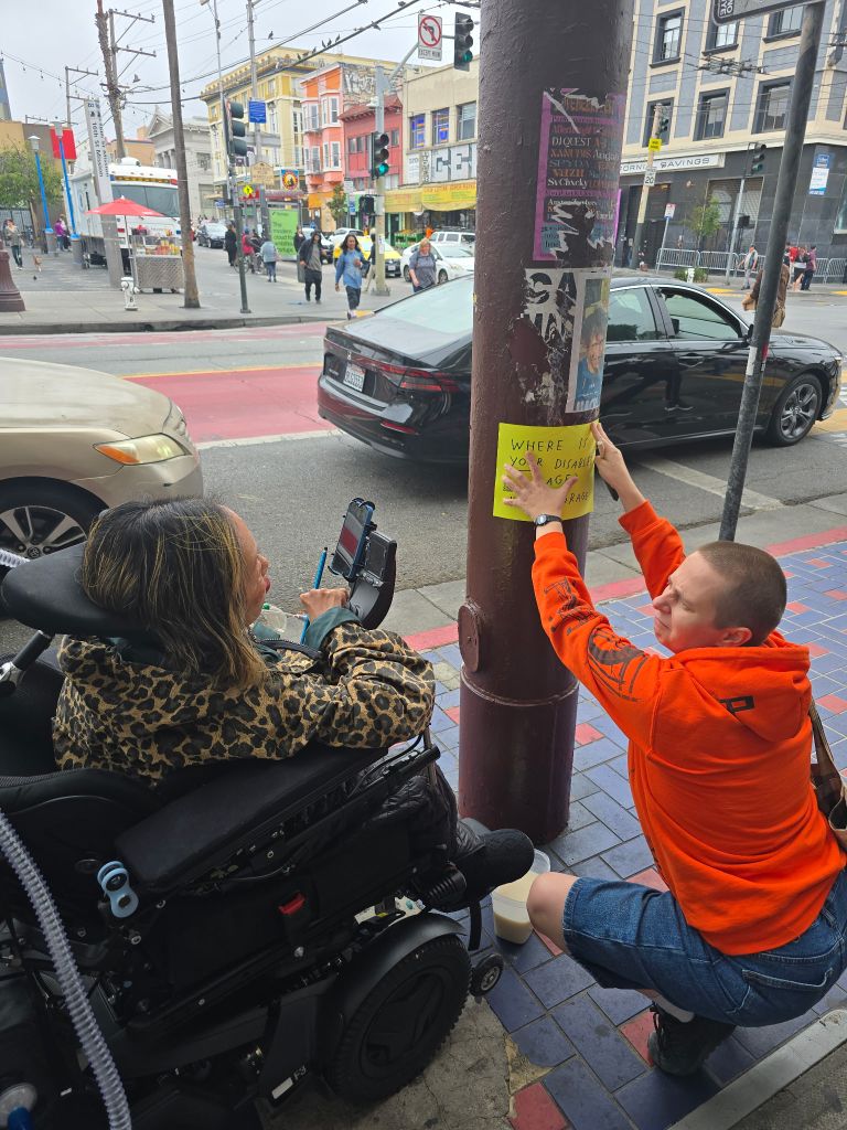 Asian American disabled woman in a wheelchair on the left and a white person on the right. The white person is knelt down pasting a bright yellow piece of paper with the text, “Where is YOUR disabled rage?”