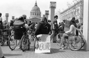 A black and white image of a group of people protesting in front of San Francisco's City Hall. Many are wheelchair users. One person's wheelchair has a sign that reads: "We shall overcome."