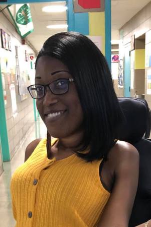 Dayniah Manderson, a young Black woman with long black hair and eyeglasses. She is in a power chair and wearing a yellow sleeveless shirt. She at her classroom door with the hallway in the background.