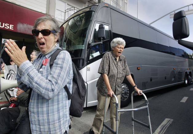 Two older adults blocking a large bus. One older woman is clapping her hand and another woman is using a walker.