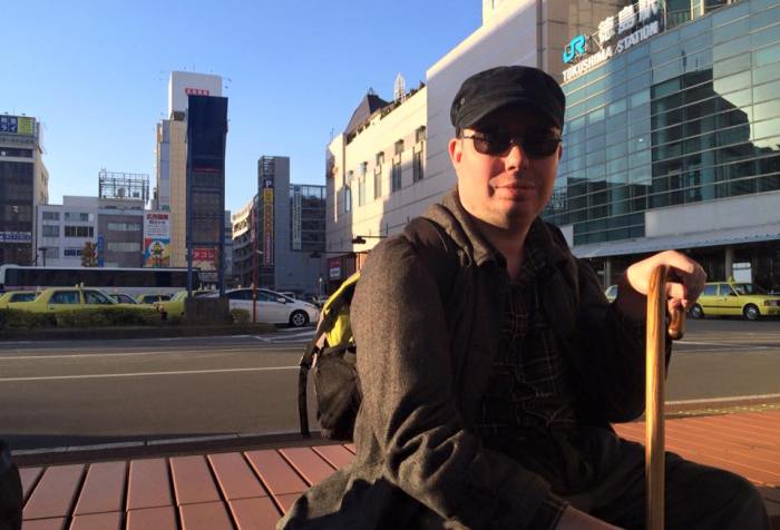 A white man sitting on a bench with a cane. He is wearing dark sunglasses and has a black cap on his head. Behind him is an urban landscape of a city in Japan