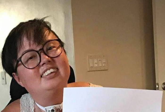 A photo of Stacey Park Milbern, a mixed race Korean and white queer person, holding a paper sign that reads “Cherished.” The photo is stylistically torn at the edges with cursive text appearing across the bottom, “Stacey Milbern.”