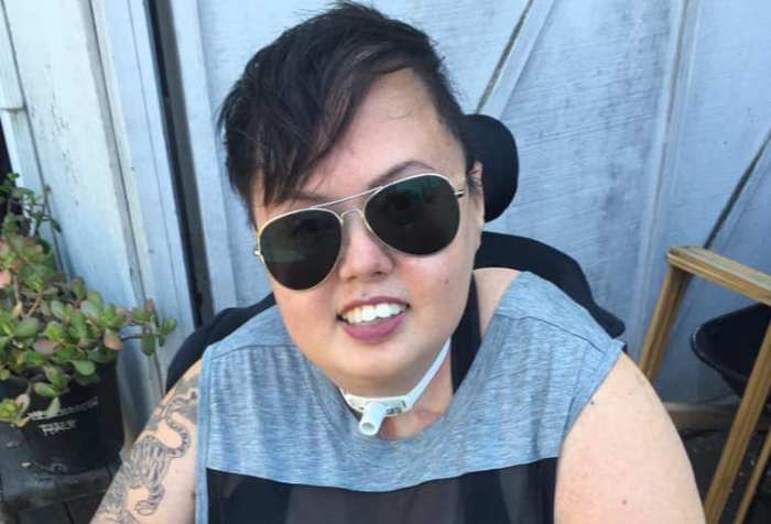 Stacey Park Milbern outside smiling at the camera with gold rimmed aviator sunglasses on. Her short, undercut styled hair is swooped forward over her forehead a little to the right. A tiger tattoo curls up her bare right arm. She is wearing a grey sleeveless tee shirt with a bold black horizontal stripe and red and black plaid leggings.