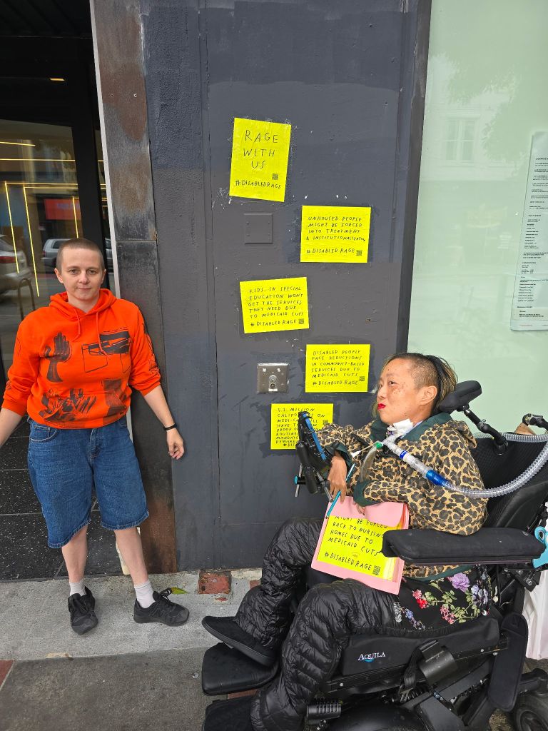 Asian American disabled woman in a wheelchair on the right and a white person on the left. In between us is a wall with a vertical row of 4 signs in bright yellow paper about the devastating impacts of Medicaid cuts on disabled people and other harmful actions by the President.