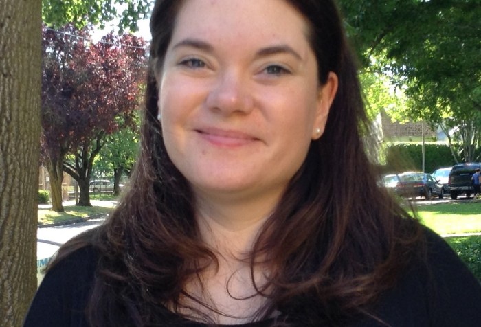 Image description: A woman with long brown hair, a black shirt, and a name tag written in Sharpie on her shirt stands outside near the trees, smiling at the camera.]