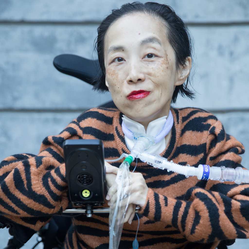 Photo of Alice Wong, an Asian American disabled woman in a power chair. She is wearing a black blouse with a floral print, a bold red lip color and a trach at her neck. She is giving a cheeky expression with her eyebrow partially raised. In the background is a gray cement wall. Photo credit: Eddie Hernandez Photography.