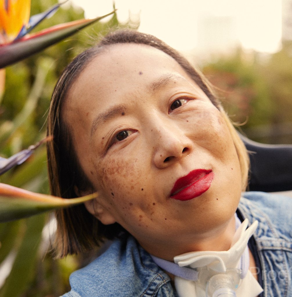 Alice Wong, an Asian American disabled woman with a tracheostomy at her neck. She is wearing a bright red lip color and a denim shirt. She is smiling with her head tilted toward some bird of paradise plants. Photo credit: María del Río