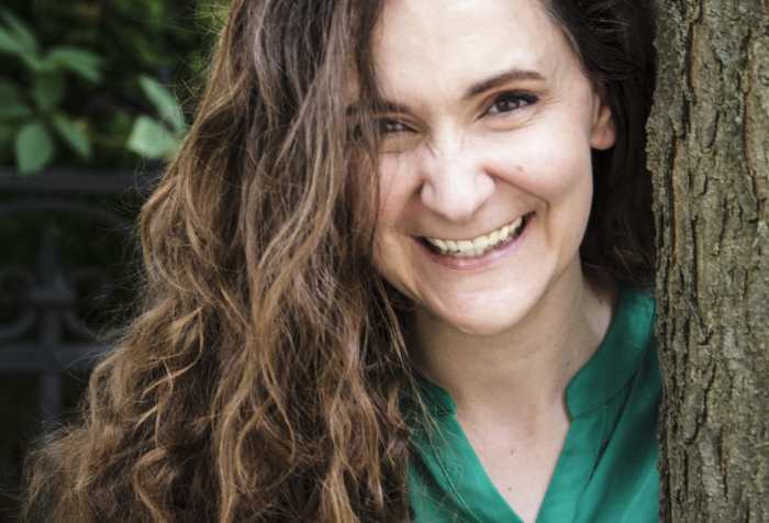 A brown-haired brown-eyed woman stands smiling beside a tree trunk. She wears a deep green shirt and an ivory cardigan.
