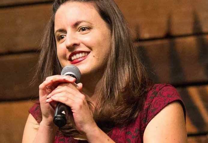 Nina G, a young white Italian American woman with long brown hair and a short-sleeved red dress smiling and holding a microphone.