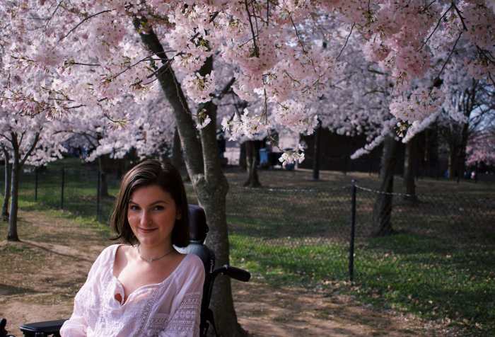 A young white woman with chin-length brown hair smiles at the camera. She wears a white shirt and sits in a black motorized wheelchair. Behind her are several blooming cherry blossom trees.