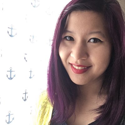 Photo of Elea Chang, a smiling Asian woman with shoulder-length purple hair and a yellow cardigan looking directly at the camera. She is to the right of the frame, in front of a white curtain patterned with blue anchors.