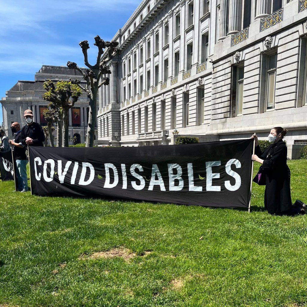 Two people in black clothing and wearing n-95 respirators hold a large black banner with white all-caps text that reads “covid disables”. They are in a green grassy area next to the ornately adorned SF City Hall. Two people are just to the left of them, you can’t read the banner they are holding, but it is black with white text and it says “covid kills”.