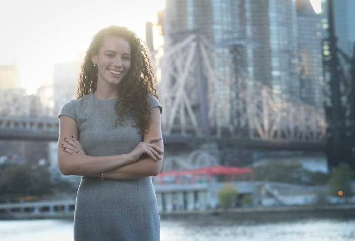 Rebecca Lamorte, candidate for New York City Council, is smiling and standing on Roosevelt Island in New York City in front of the Queensboro Bridge. Rebecca is a white woman with long, curly brown hair and is wearing a gray dress. 
