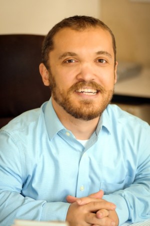 Short statured caucasian male in his late 30s sitting in a black chair and smiling with his arms crossed. He is wearing a blue collared shirt.