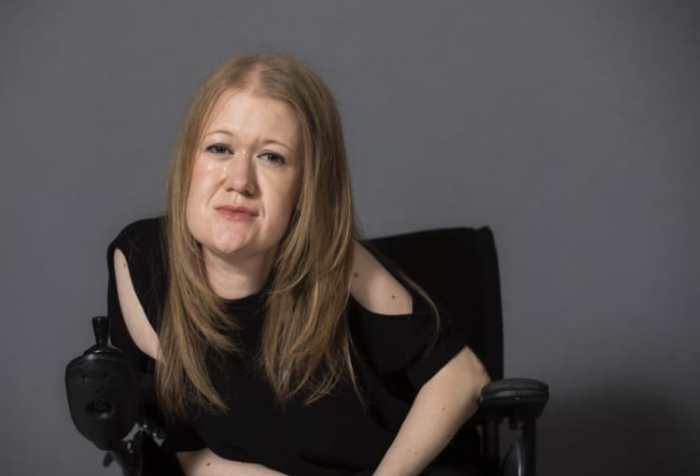 Picture is of Frances sitting in her wheelchair; smiling, wearing a black top and her blonde hair down.