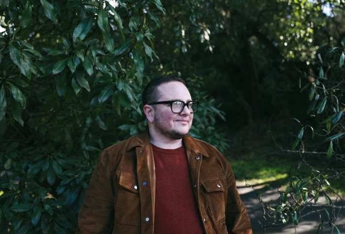 A white non-binary person smiles with trees and green plants in the background.