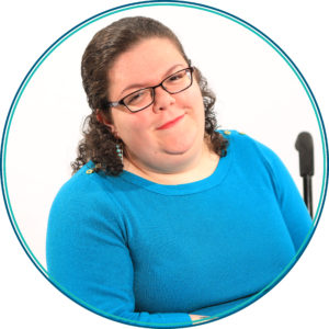 Image of Emily Ladau a young white woman with curly brown hair. She is wearing glasses and a blue sweater. She is sitting in a wheelchair.