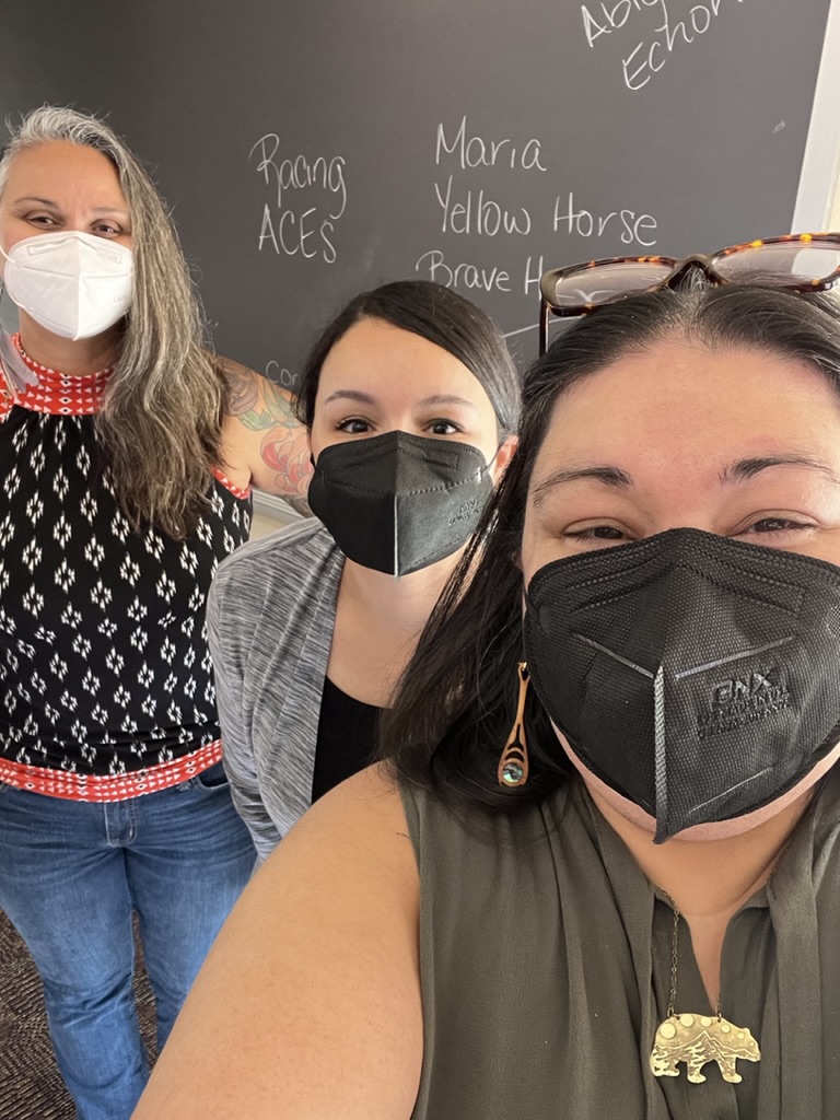A photo of three UPRISE COLLECTIVE staff members. Brianna who is on the right is a Native/Ihanktonwan person, wearing black mask, green shirt, golden bear-shaped necklace, and wood earrings with abalone shells. In the middle is Stephanie, a Black/Native woman wearing black mask and grey cardigan. Their black hair is tied in the back. On the left is Ginia, a Blauan person, wearing a white mask, multi-colored shirt, and jeans. They are all posing in front of a blackboard.