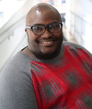 Photo of Finn Gardiner, a bald black man in his 30s wearing glasses and a red and grey sweater. 