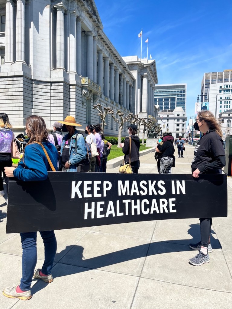 Two people, facing to the left of the camera, are holding a black sign with white writing that says “Keep masks in healthcare”. In front of them is a crowd of people also faced to the left of the camera, at the corner of San Francisco's column enshrined City Hall.