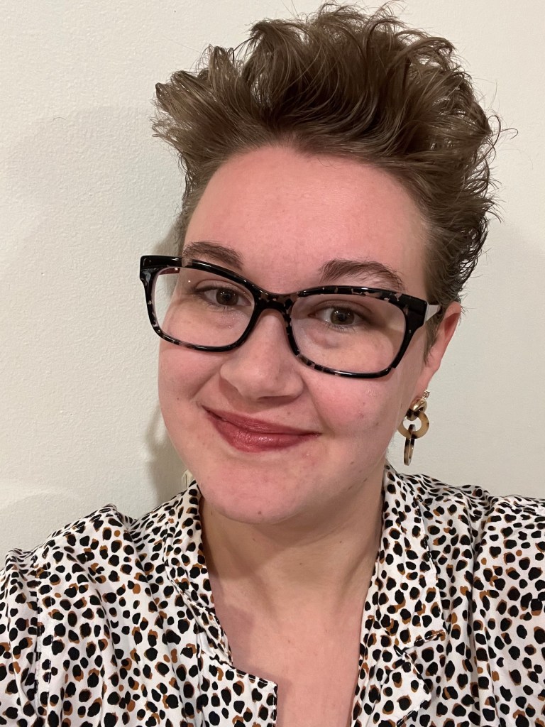 photo of Helen, a white person with short brown hair and glasses wearing a patterned shirt and red lipstick