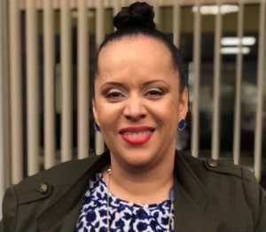 A light-complected Black woman shown shoulders up wearing hair in a bun atop her head, blue button earrings, makeup with red lipstick and smiling. She is wearing olive-colored blazer and blue and white patterned blouse with long necklace of various blue-colored pendants.