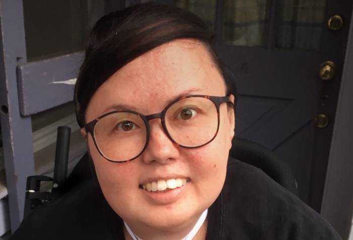 A mixed race Korean and white queer person smiles head on at the camera. She has big glasses and is wearing a crewneck. Her trach and wheelchair can be seen.