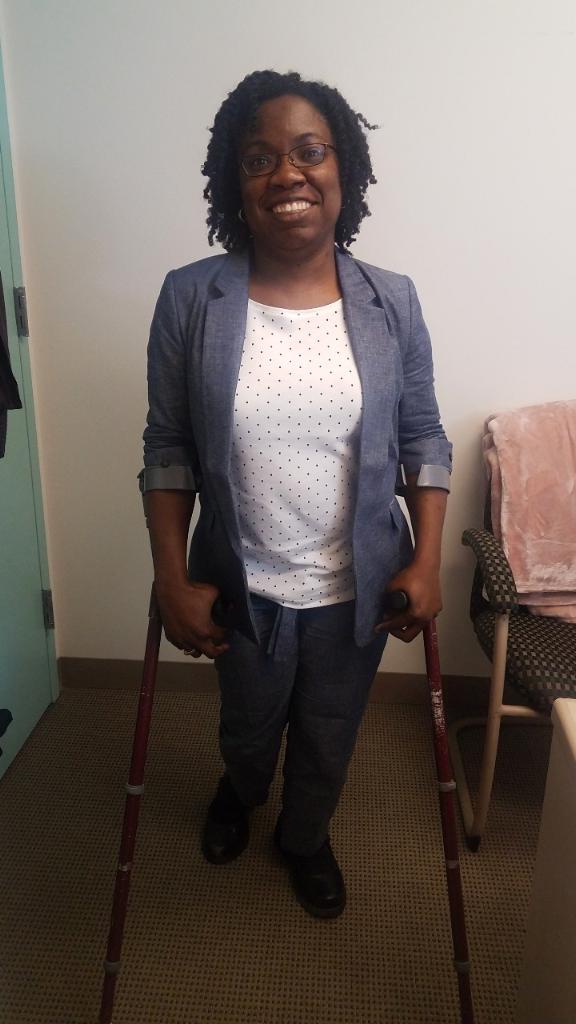 Britney Wilson, Black woman with curly natural, black hair and glasses in a blue chambray suit and white and blue polka dot top standing on crutches in an office.