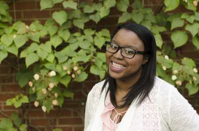 Black woman with black hair and black glasses is wearing a white shirt and smiling in front of a green flowered wall. Photo credit: Reid Beels