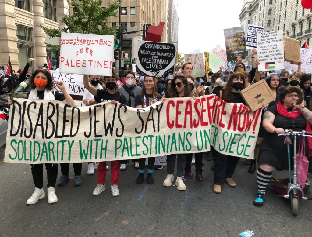 Disabled masked and unmasked jews marching in Washing DC, holding a big sign that says "DISABLED JEWS SAY CEASEFIRE NOTE! SOLIDARITY WITH PALESTINIANS UNDER SIEGE". Disabled Jews, include Nomy Lamm on a scooter holding a handmade shofar, wearing a red scarf and black dress with a visible ribcage pattern.  There are other signs in the background that include "Free Palestine" and "Grandchild of Holocaust survivors for Palestinian Lives" Photo Credit: Emily Schuman