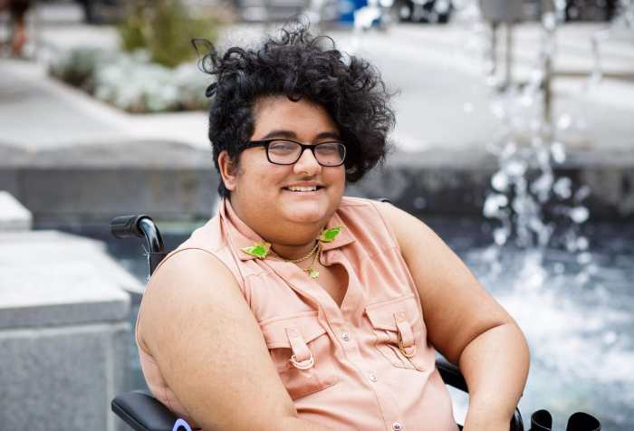 Noor sits in a wheelchair in front of a fountain. He has a curly black undercut and medium golden tan skin. He wears thick glasses, and a sleeveless pink shirt with a set of budgie collar pins. Photo credit: Les Talusan photography
