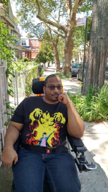 Photo of André J. Daughtry, a Black man sitting in a motorized wheelchair, wearing a black T-shirt and blue jeans.