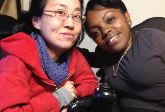 A photo featuring Yomi Wrong and Alice Wong taken on April 11, 2015. On the left is an Asian American woman in a wheelchair wearing a red hoodie and purple scarf. She has black hair, eyeglasses on, and a smartphone in her hand. On the right is a Black woman with short hair who is in a wheelchair. She is wearing a long-sleeved black shirt and a gold necklace. Both are smiling at the camera.