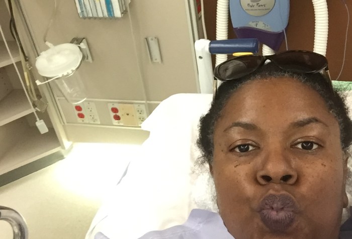 Black woman taking a selfie from her hospital bed. She has a blanket wrapped around her up to her neck and behind her are various medical devices and equipment on the wall. She is puckering her lips and looking right at the camera.