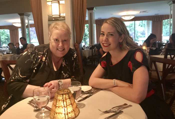 Two women with blonde hair smiling and posing for the camera at a table at a fancy restaurant in Harrison Hot Springs. On the left is Cathleen wearing a dark flower printed blouse and on the right is Amanda, the writer, wearing a black dress with roses on the sleeves. They look happy and rested.