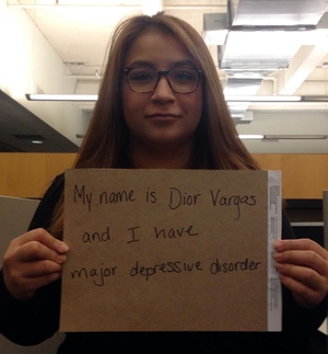 Photo of a Latina with long brown hair and wearing glasses. She is holding a sign that reads: "My name is Dior Vargas and I have major depressive order."