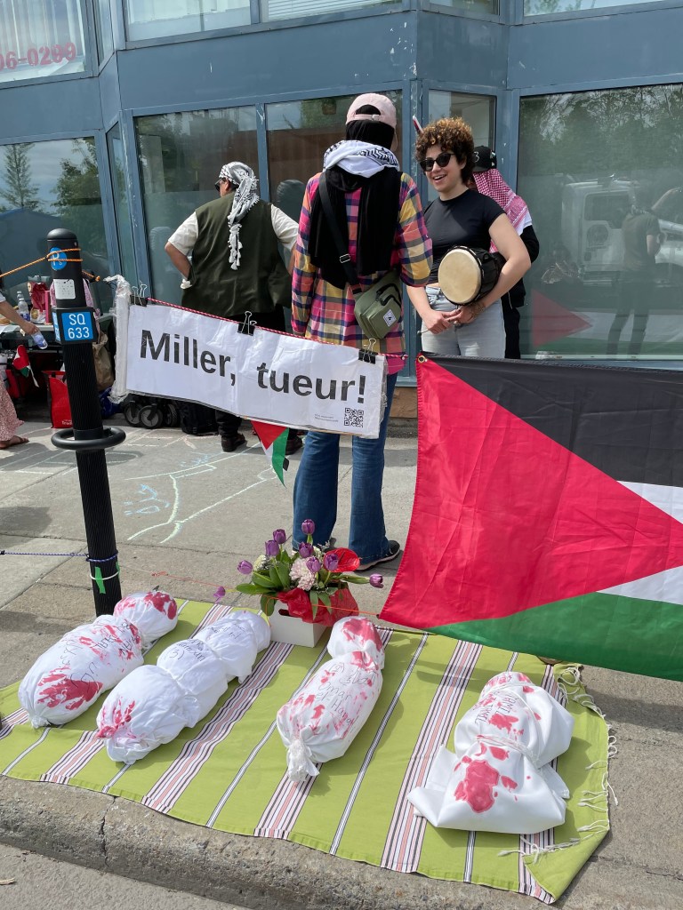 Photo of Samar AlKhdour wearing blue jeans, a colourful checkered shirt, a black and white keffiyeh and a light pink baseball cap in conversation with a sit-in supporter in front of Canadian Minister of Immigration Marc Miller’s riding office in Montreal on May 17, 2024. Behind them are the Immigration minister’s outside office-building walls where all the posters and stickers in support of Palestinian liberation were removed. Samar received orders not to allow more postering on the walls or face fines and charges. In the foreground, a Palestinian flag hangs on a rope next to a white paper that has been clipped onto that same rope that reads in French: “Miller, Tueur” (which translates to Miller, Killer). In front of that, a makeshift graveyard of four ‘killed babies’ wrapped in white cloth with bloodstains (replicating the way killed Palestinian babies are shrouded in Gaza for burial). Photo by Nelly Bassily