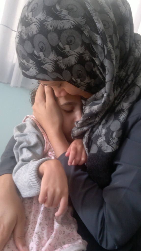 Samar Alkhdour, a Palestinian mother who is wearing a dark gray headscarf and a black shirt. She is cradling her disabled daughter Jana. Samar's head is bent down, her arms cradling Jana.