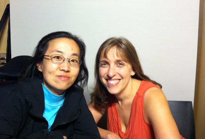 Asian American woman on the left side in a wheelchair, she is wearing glasses and wearing a black hoodie. On her right is a white woman with long blonde-brown hair wearing an orange tank top. Both are smiling at the camera.